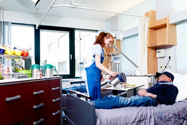 Serviceassistent staat aan het bed van een patiënt en schenkt koffie in Serviceassistent staat aan het bed van een patiënt en schenkt koffie in