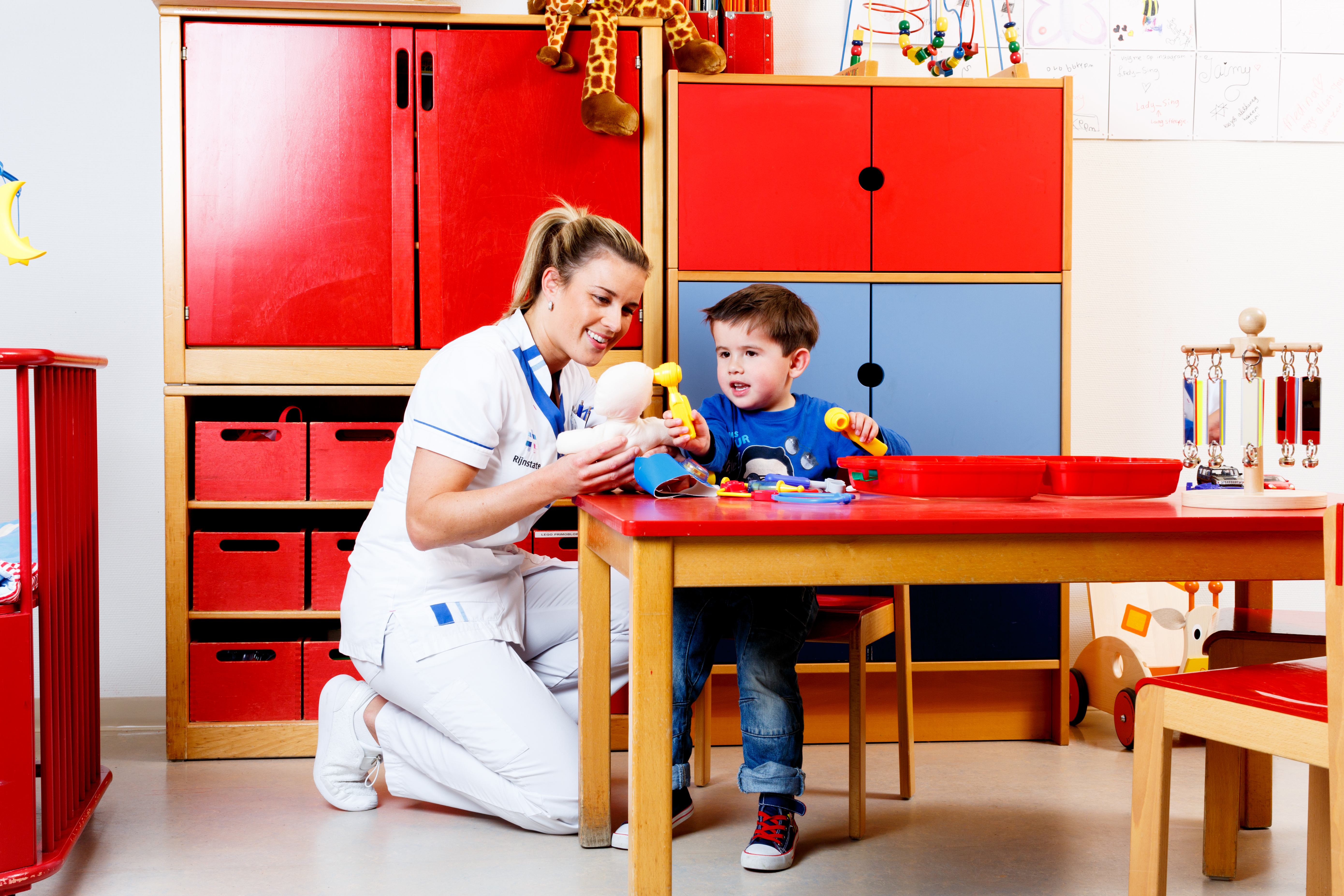 Verpleegkundige zit naast een kind dat met een oorspiegel in de oren van een pop kijkt