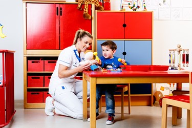 Verpleegkundige zit naast een kind dat met een oorspiegel in de oren van een pop kijkt Verpleegkundige zit naast een kind dat met een oorspiegel in de oren van een pop kijkt